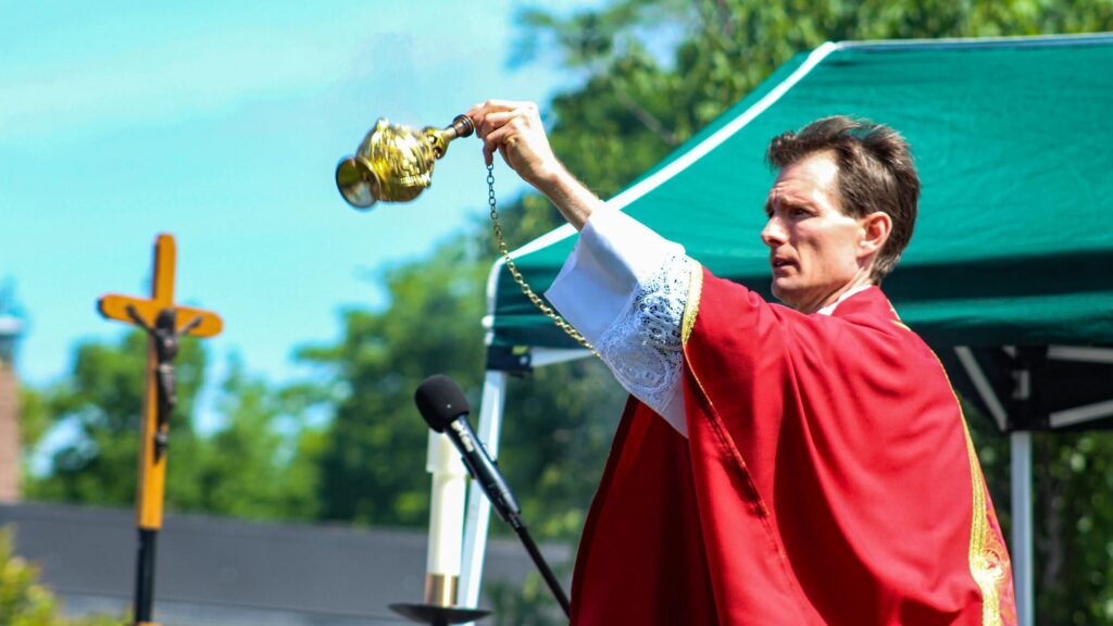 Priest in red vestments holding an ornate golden censer during an outdoor liturgy