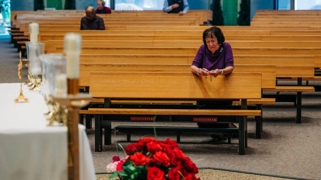 Woman praying alone in a church pew beside flowers and candles